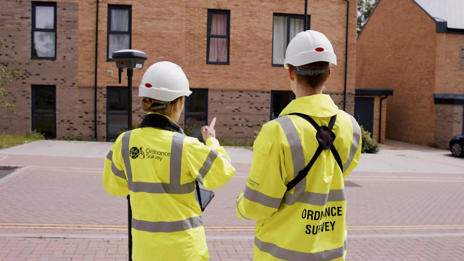 Two OS surveyors carrying GNSS equipment and wearing OS branded high-visibility jackets.