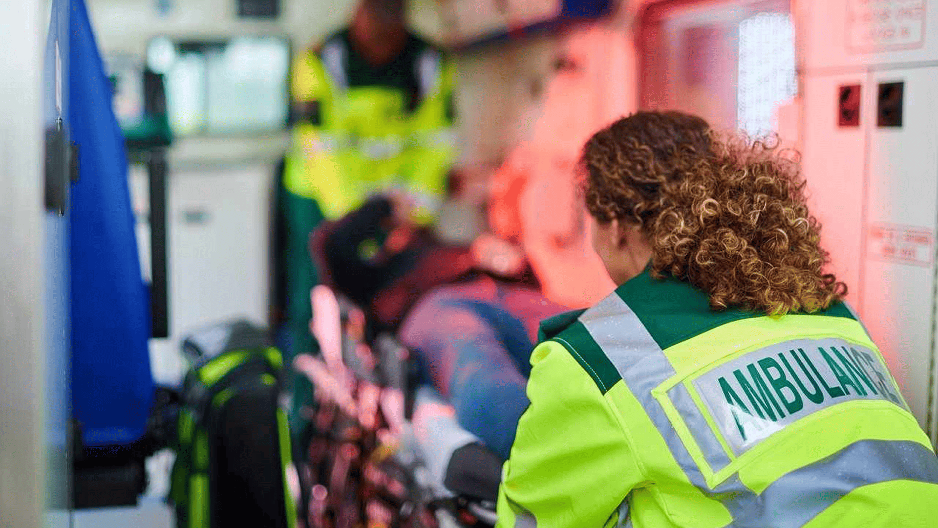 A woman with curly hair wearing a high vis jacket with the word ambulance on the back