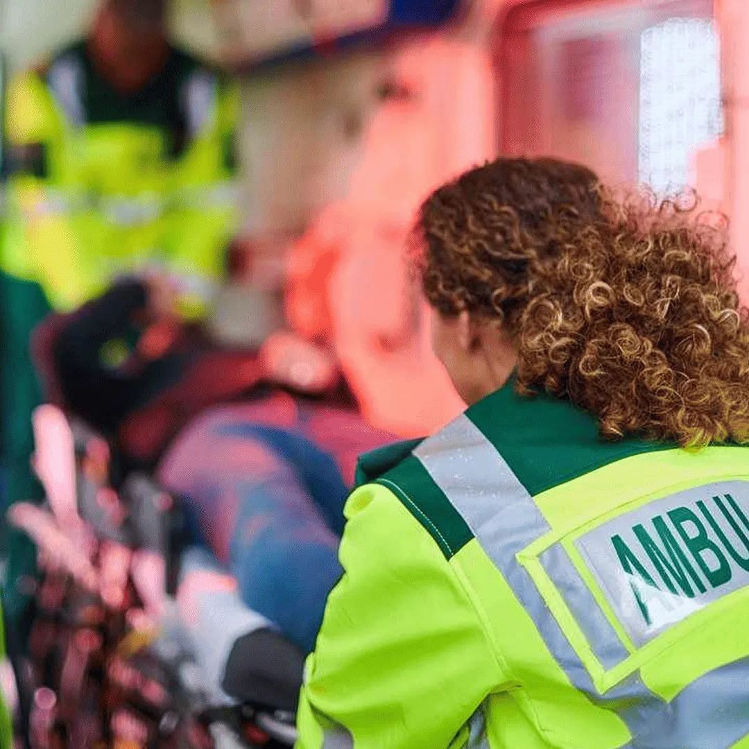 A woman with curly hair wearing a high vis jacket with the word ambulance on the back
